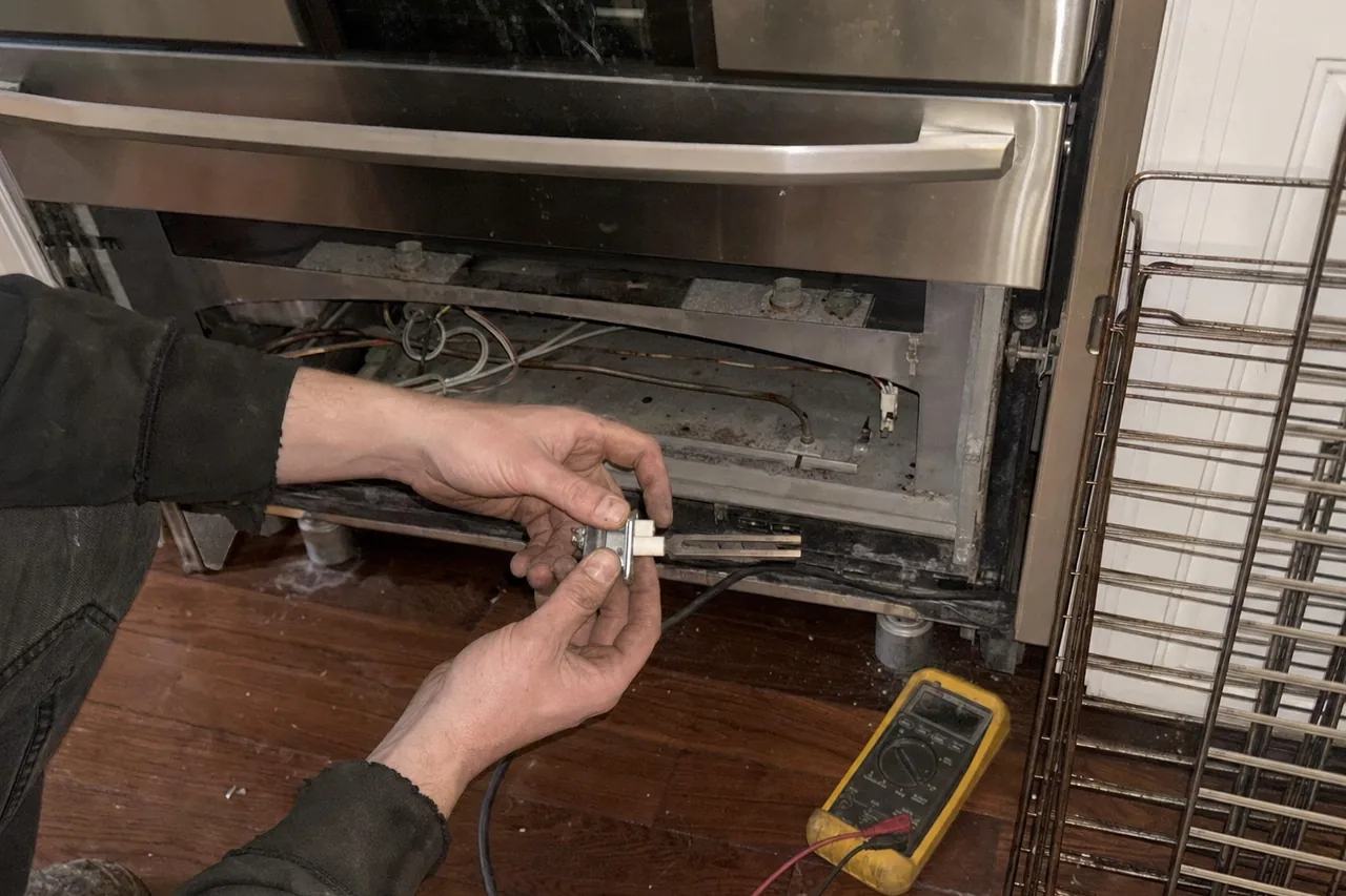Oven igniter and its ceramic mount held in front of the open lower cavity of a stainless range, burner tube and loom behind