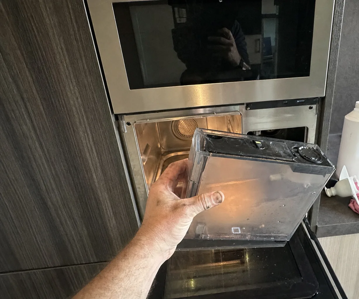 Hand withdrawing a clear water tank from a built-in steam oven, the lit cavity and its perforated steam plate visible behind