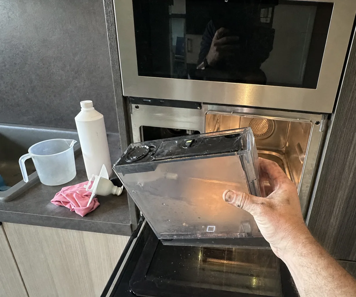 Clear water reservoir held beside its built-in appliance, with a descaling bottle, a funnel, a measuring jug and a cloth waiting on the counter