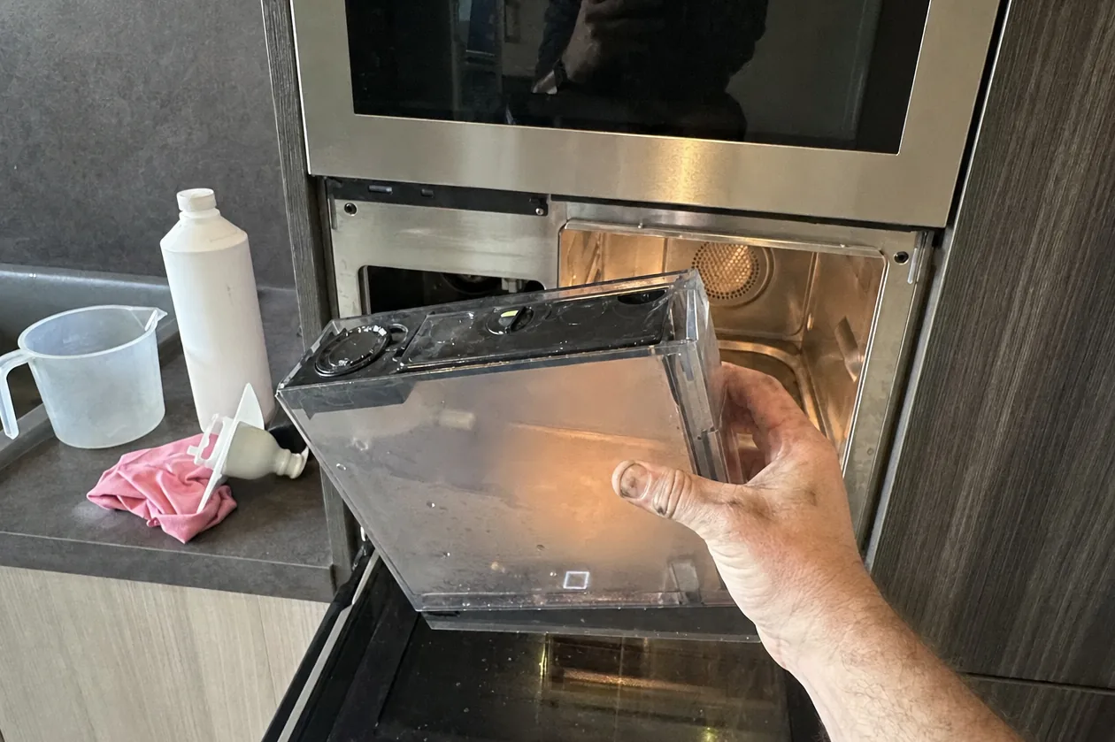 Hand drawing the clear water reservoir out of a built-in steam oven set into a run of pale cabinetry, a measuring jug and a folded cloth on the worktop beside it