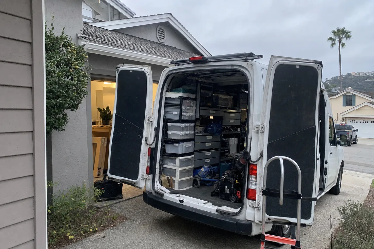 White service van backed up a residential driveway with both rear doors open, parts drawers inside and a hand truck strapped to one door